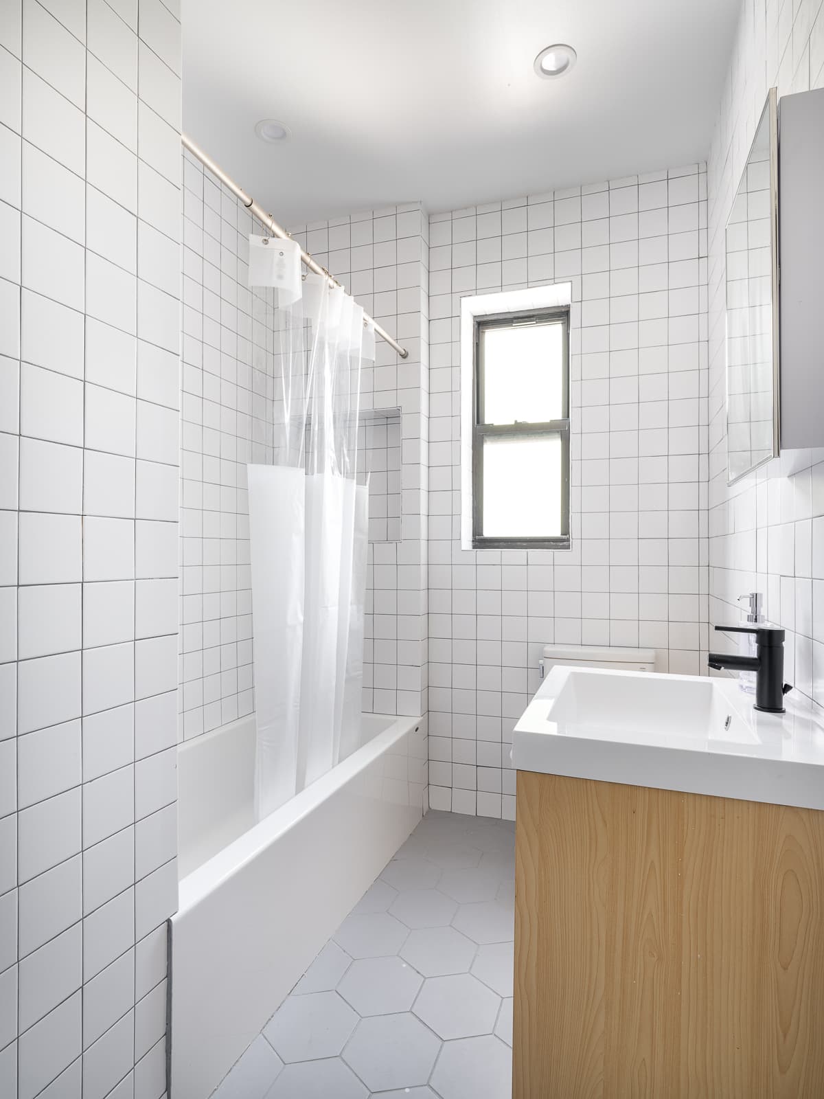 White square-tiled bathroom with bathtub, window and wood vanity