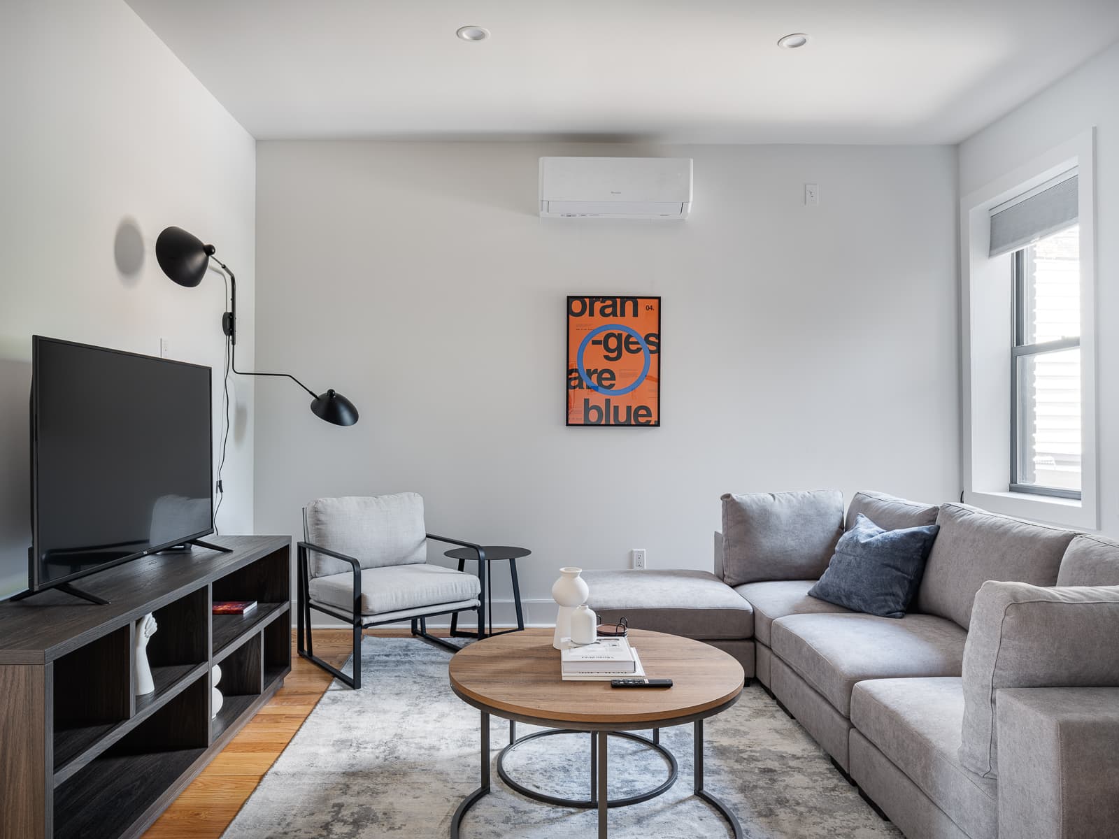 Living room with grey sectional, round wood coffee table and an orange "oranges are blue" poster